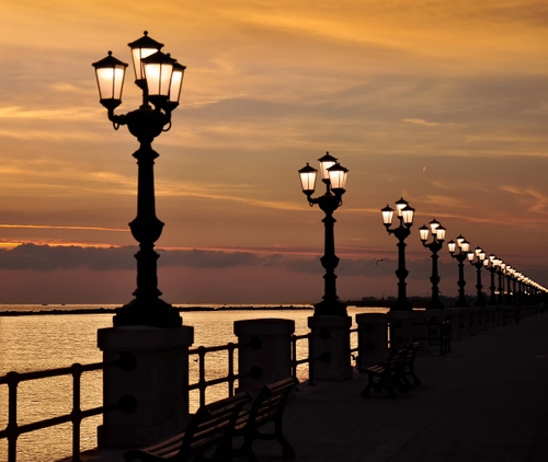 Immagine con lampioni, mare, tramonto, lungomare, panchine, nuvole, prospettiva, luci