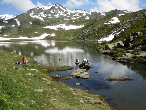 Immagine con neve, montagne, lago, riflessi, cielo, montagna, persone, nuvole, verde, erba, azzurro, rocce, acqua, riflesso, bianco, sassi, prato, massi