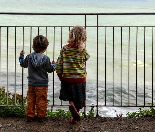 Immagine con bambini, mare, ringhiera, acqua, erba, due, onde, bambino, terra, affaccio, verde, maglie, bimbi, bambina, sbarre, gonna